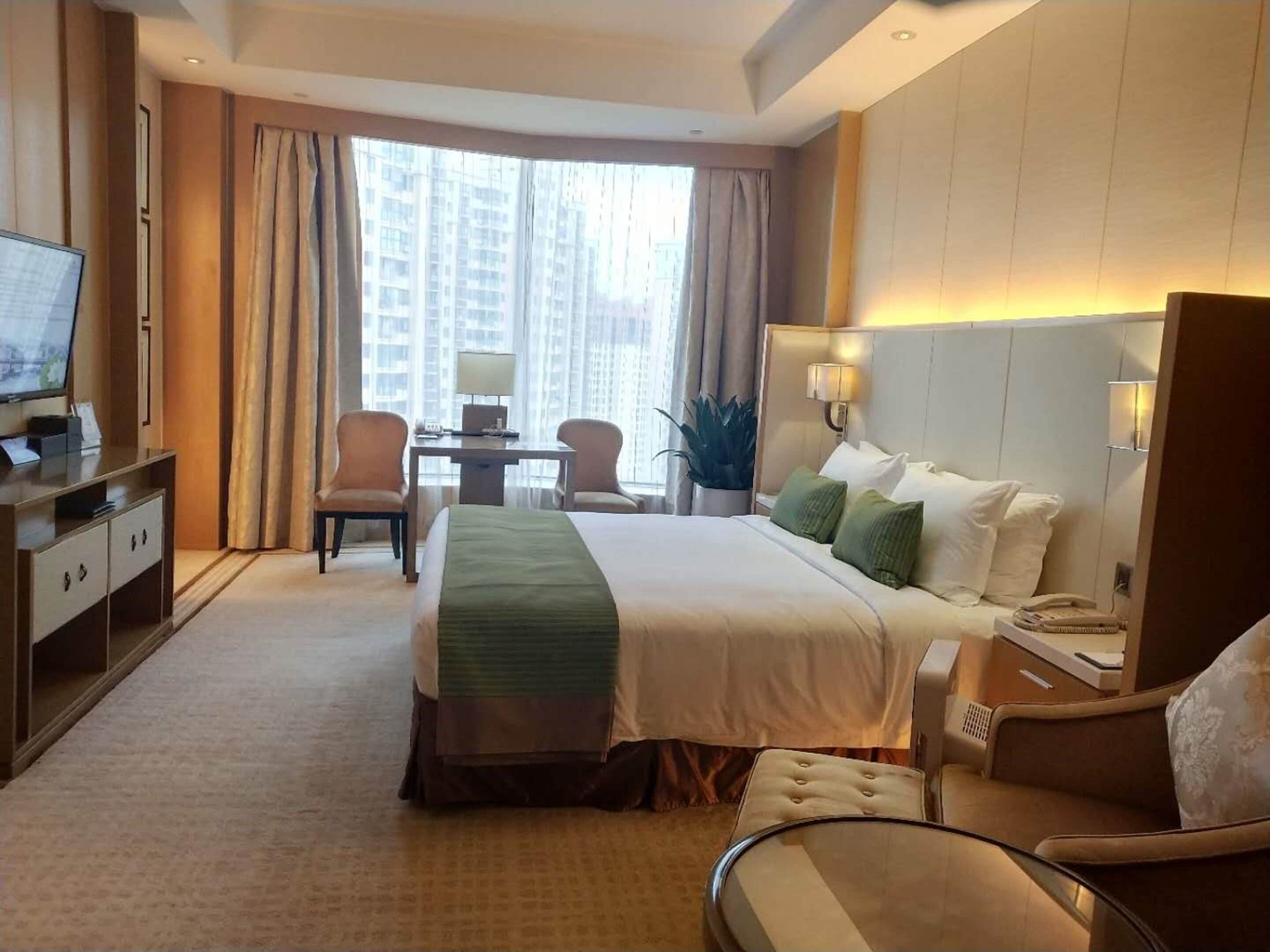 A hotel room with a large bed, a desk, a chair, a TV, and a view of high-rise buildings.