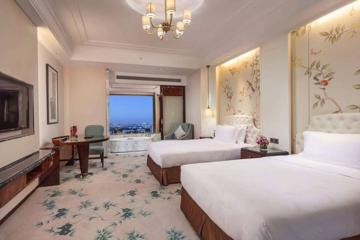 Une chambre d’hôtel avec deux lits, une télévision, un bureau avec une chaise, un lustre et une vue sur la ville.