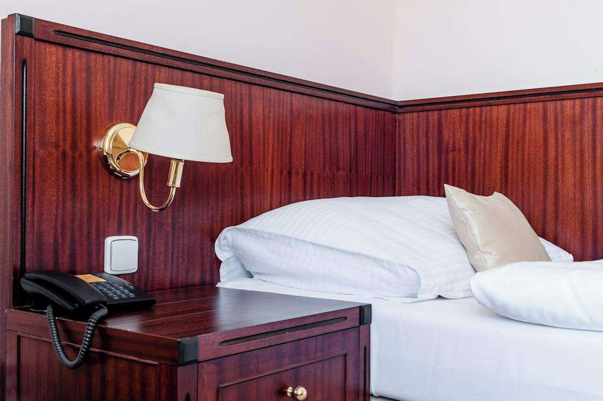 A hotel room with a wooden headboard, a bedside lamp, a telephone, and a bed with white bedding.