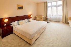 Ein Hotelzimmer mit einem großen Bett, einem Nachttisch mit Lampe, einem Sessel, einem kleinen Tisch und einem Fenster mit Vorhängen.