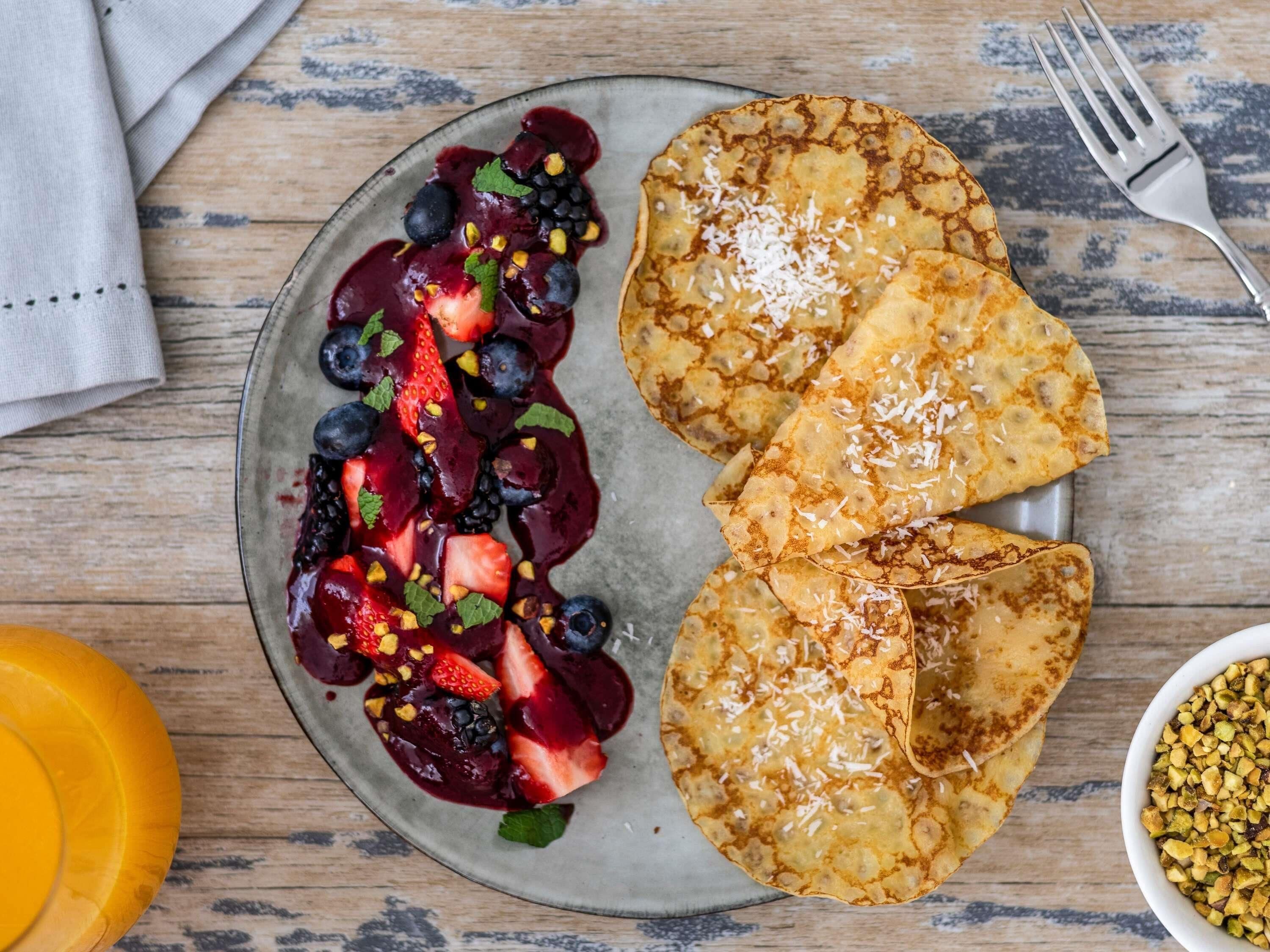 A plate of pancakes with berries and coconut flakes, a fork, and a bowl of chopped nuts.