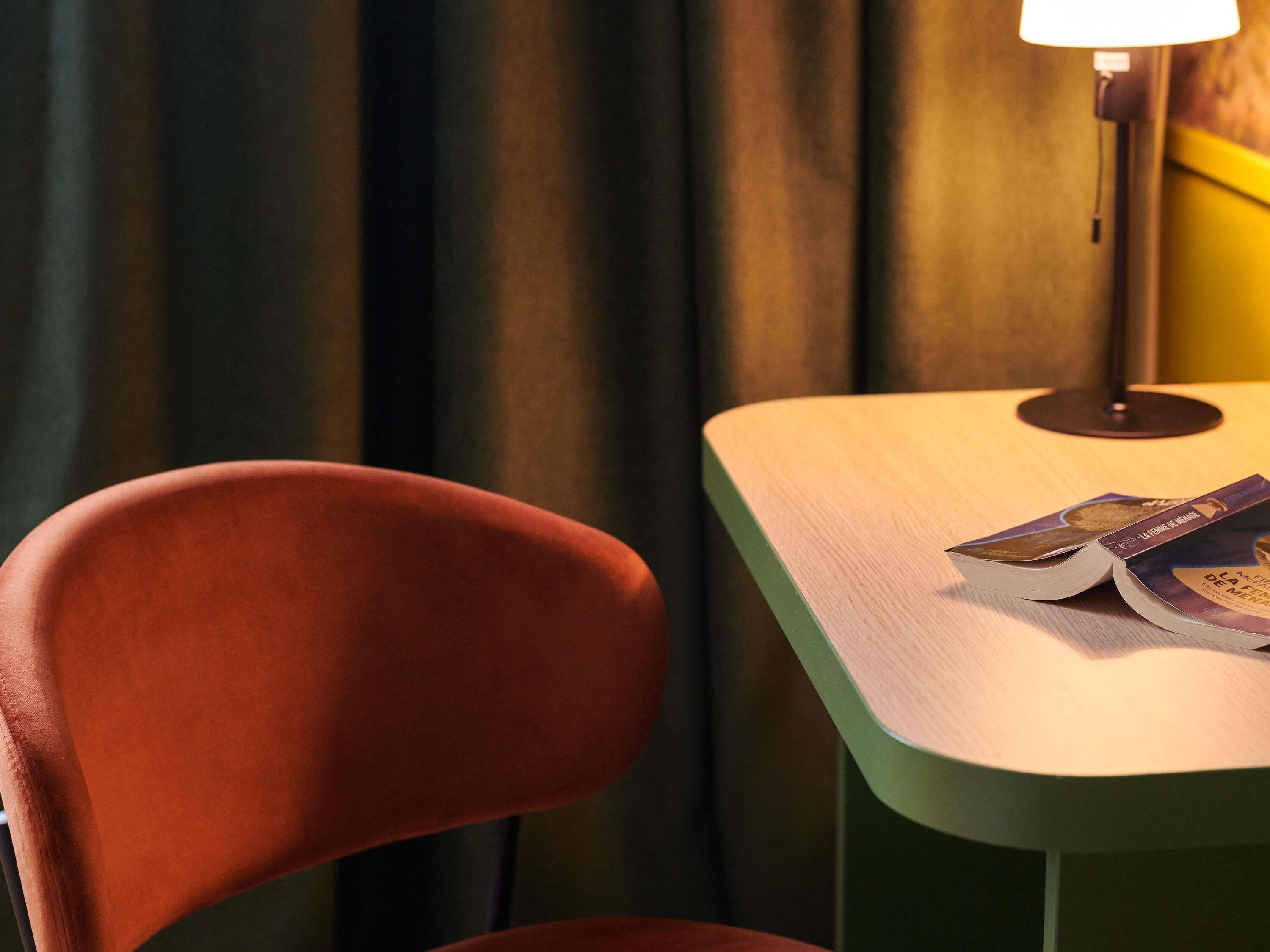 A chair and a table with a magazine, a lamp, and curtains in the background.
