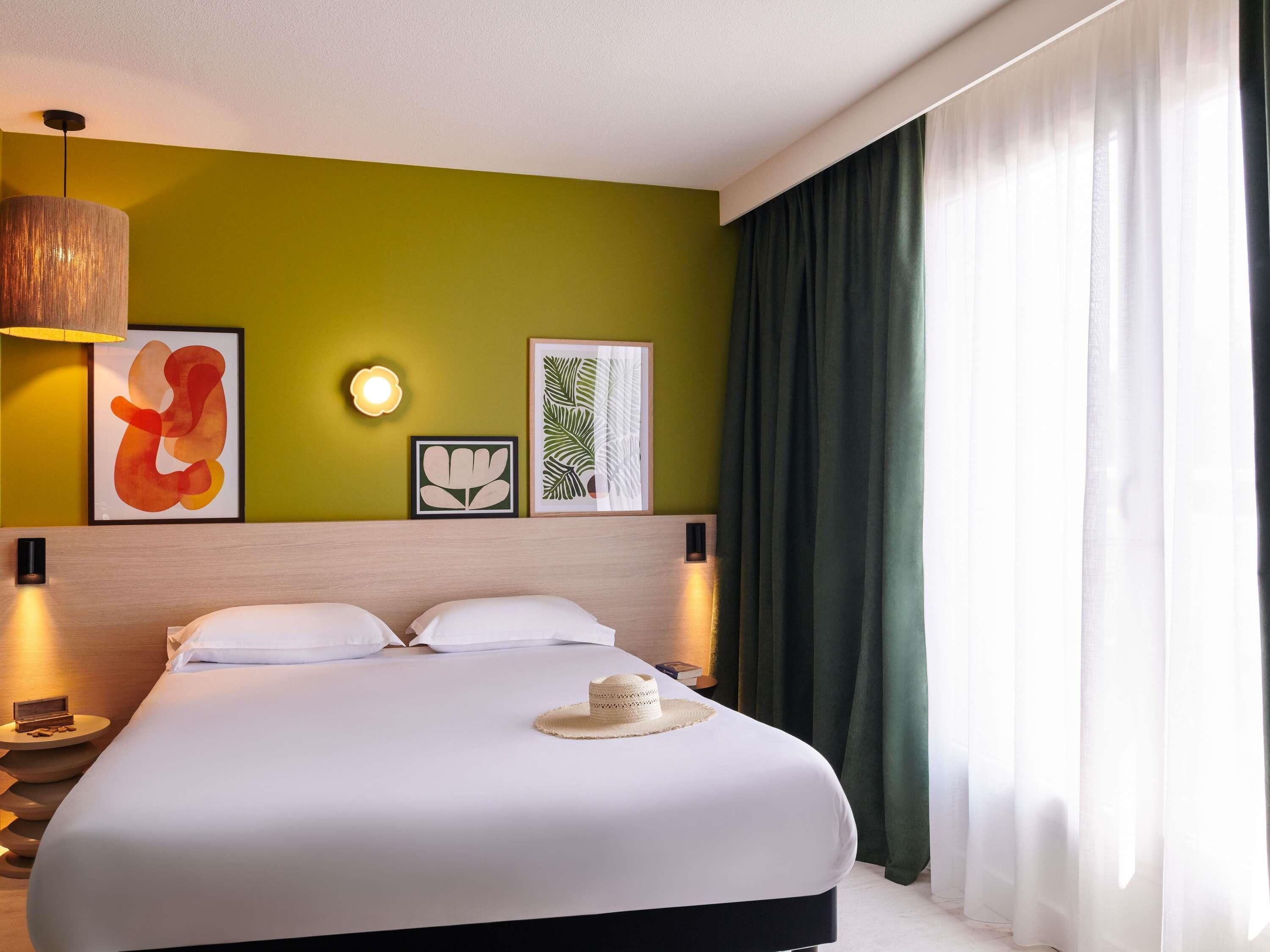 A hotel room with a large bed, a headboard, wall-mounted artwork, a bedside lamp, and a window with curtains.