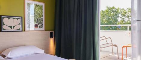 A hotel room with a bed, a window with curtains, and a balcony with chairs.