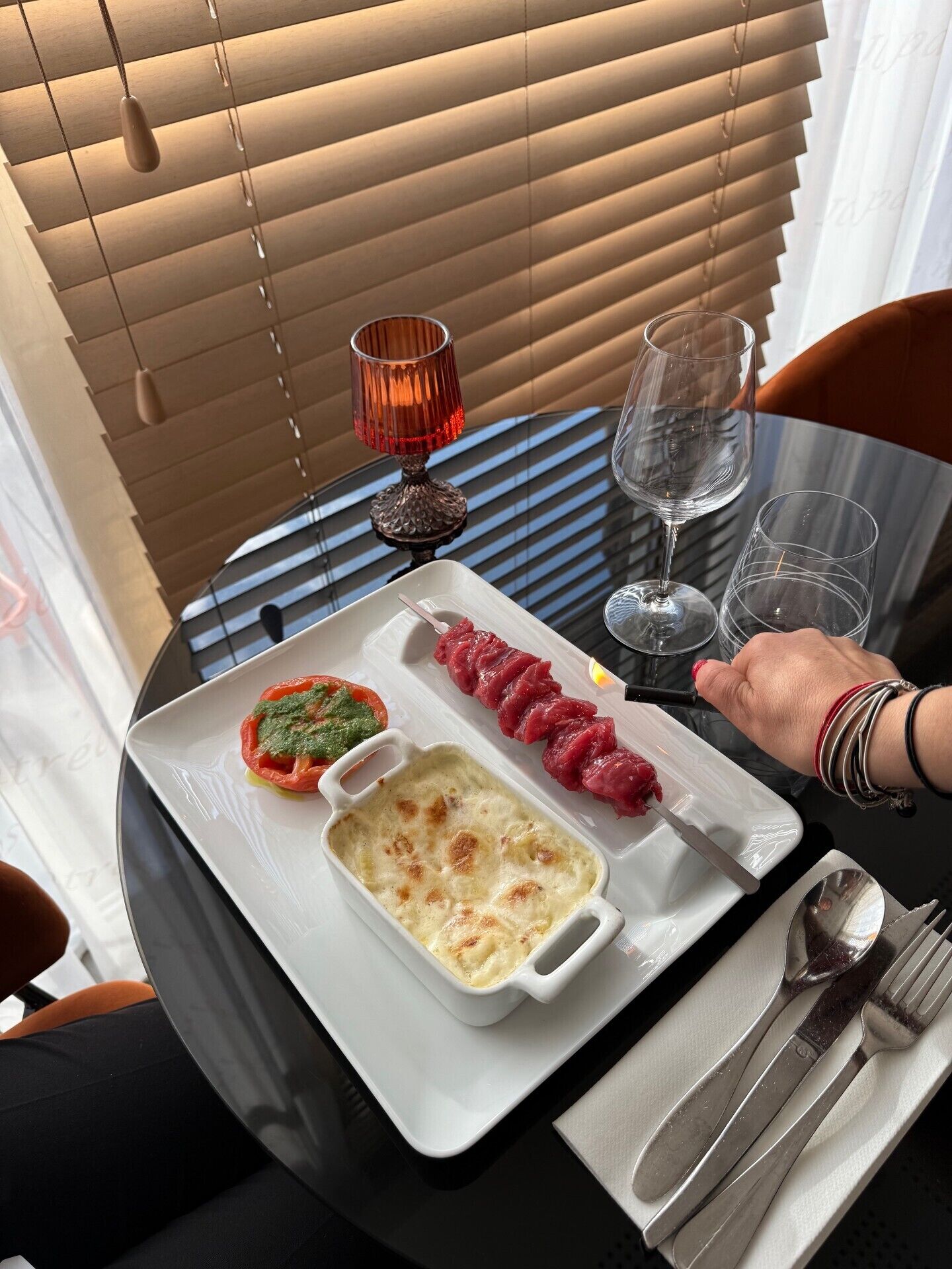 Una mesa de comedor con un plato blanco que contiene un plato al horno, un pincho de carne y un vaso de agua.