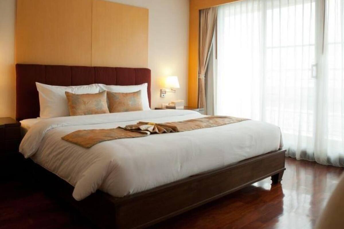 A hotel room with a large bed, a wooden headboard, a bedside table with a lamp, and a view of a building through sheer curtains.