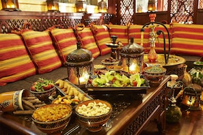 A traditional Moroccan setting with a sofa adorned with colorful cushions, a wooden table with various dishes and a hookah, and decorative lanterns. - Villa Rosa Kempinski (Nairobi)