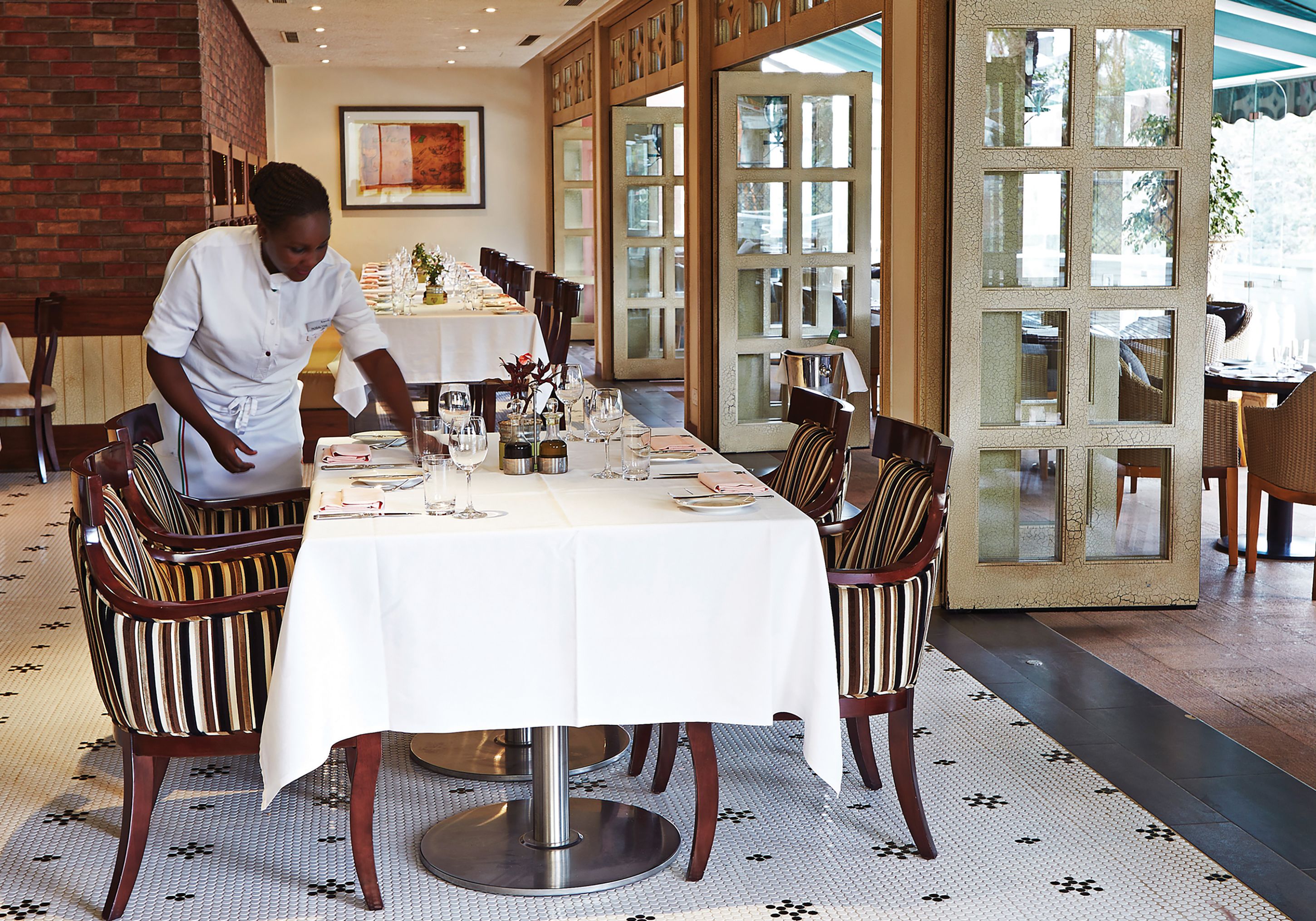 A restaurant with a chef preparing a table, featuring a patterned floor, striped chairs, and a large window.