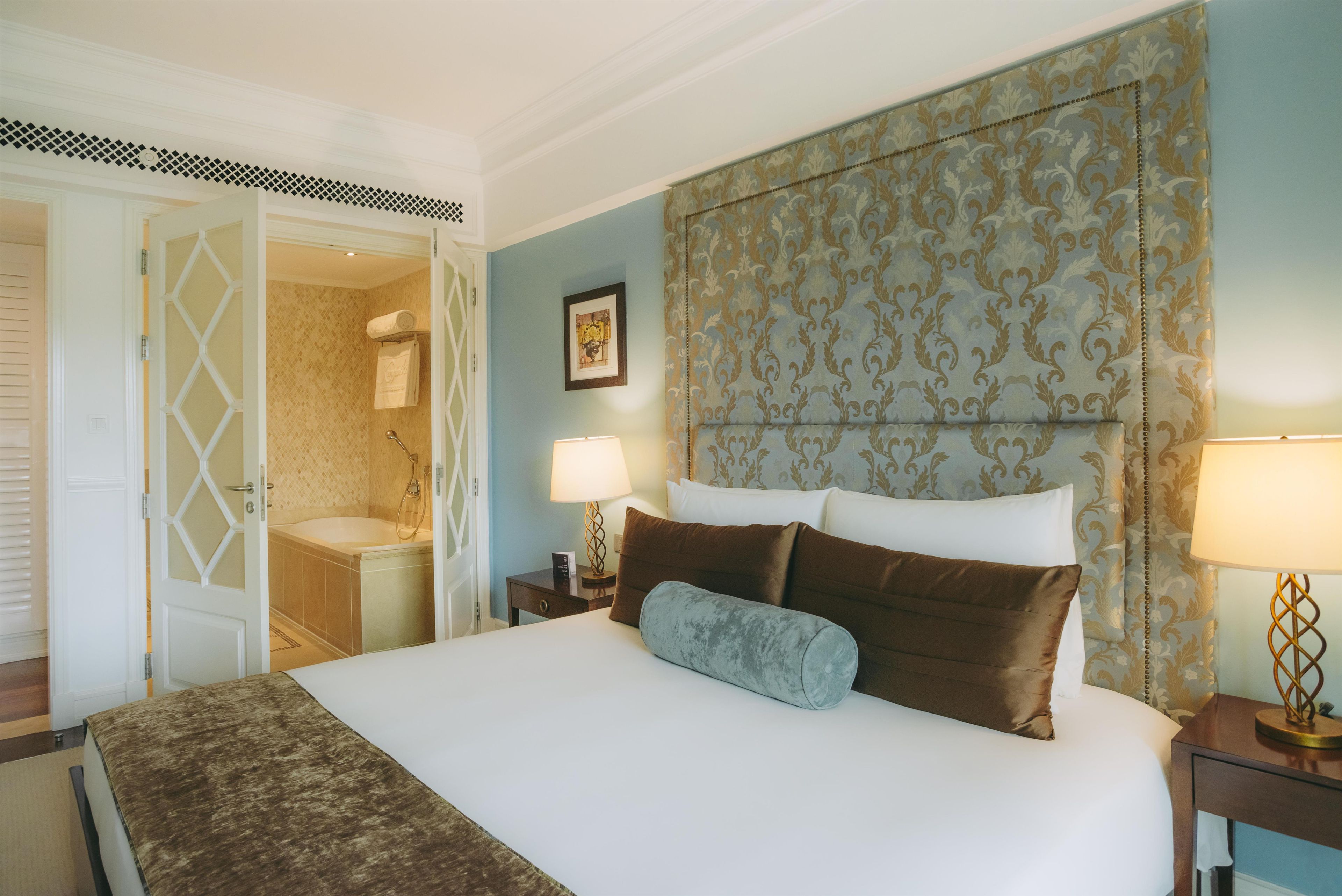 Habitación de hotel con una cama, una mesita de noche, una lámpara, un baño con ducha y una puerta que da a otra habitación.