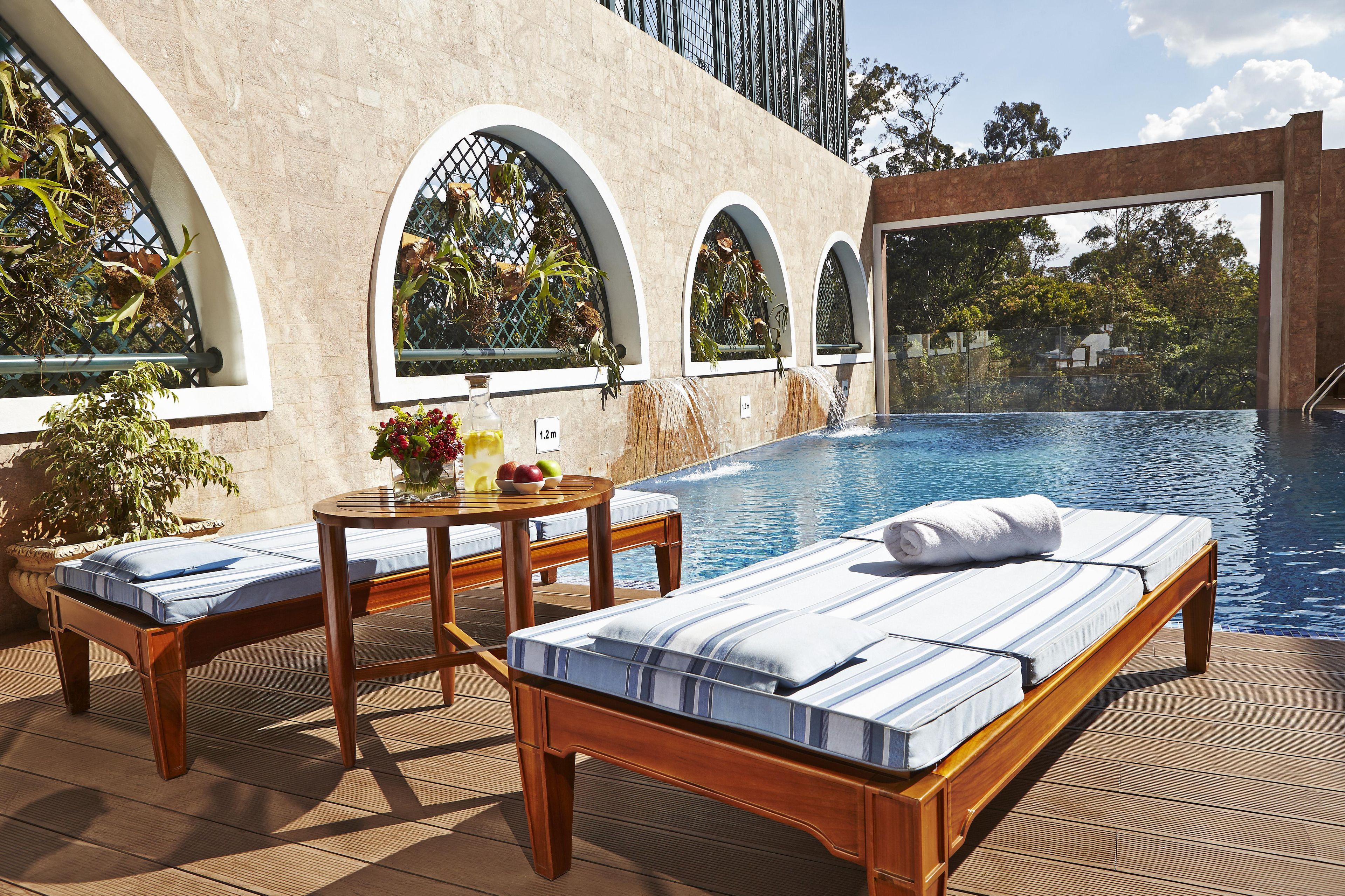 A poolside area with lounge chairs, a table with fruit and a drink, and a view of the pool and greenery.