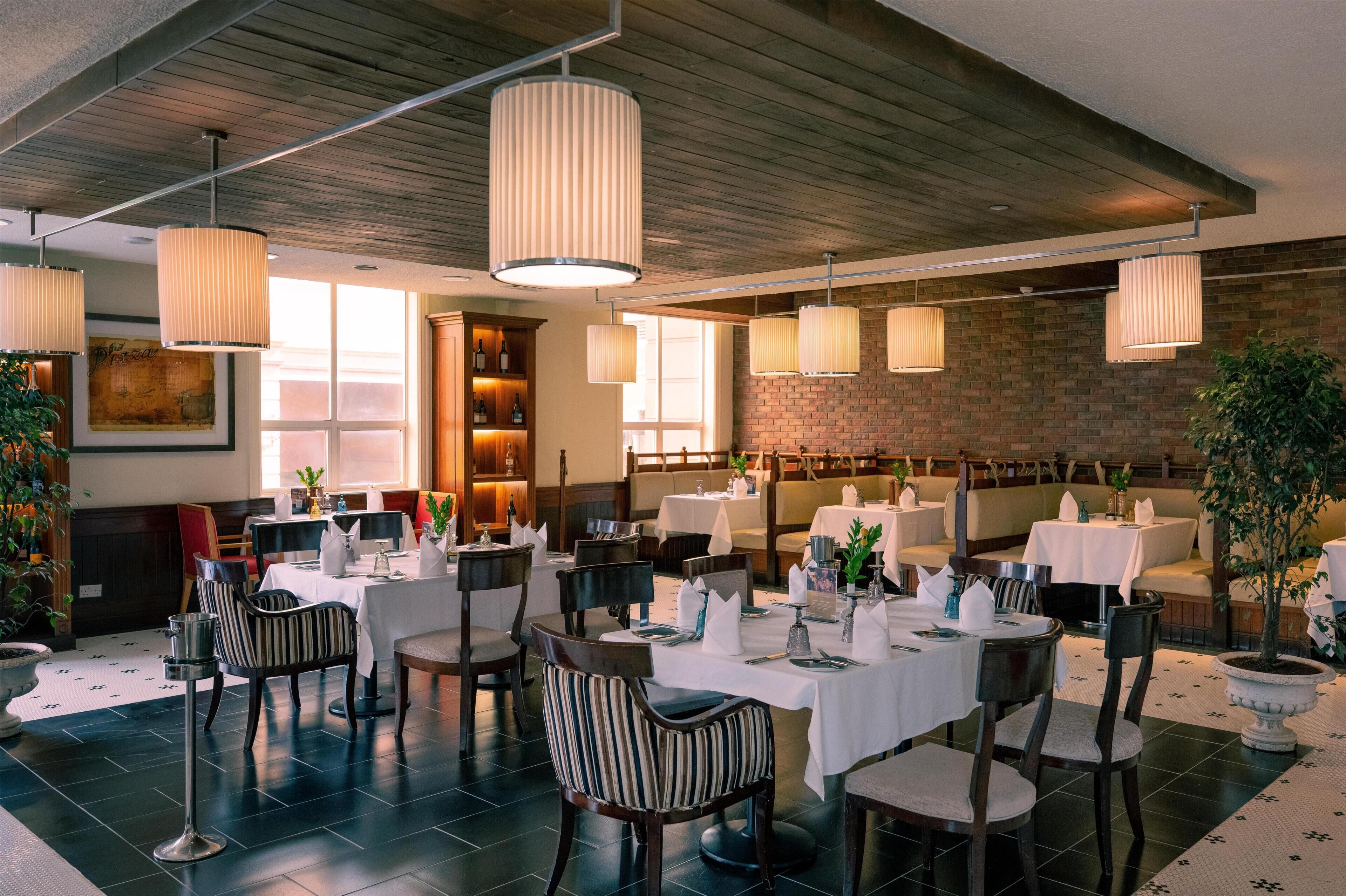 Un restaurante con mesas dispuestas para comer, lámparas colgantes, paredes de ladrillo y un amplio ventanal.