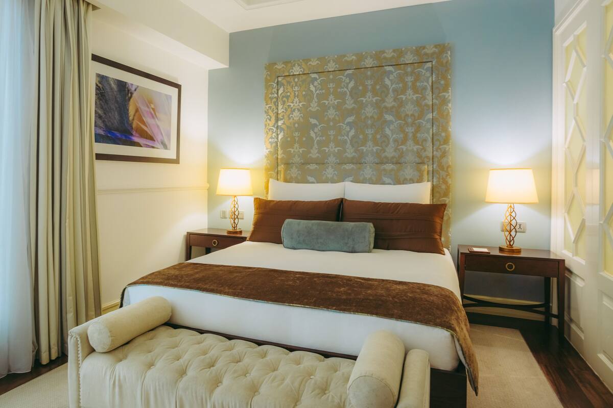 A hotel room with a large bed, two bedside tables with lamps, a headboard with a patterned design, a framed picture on the wall, and a window with curtains.