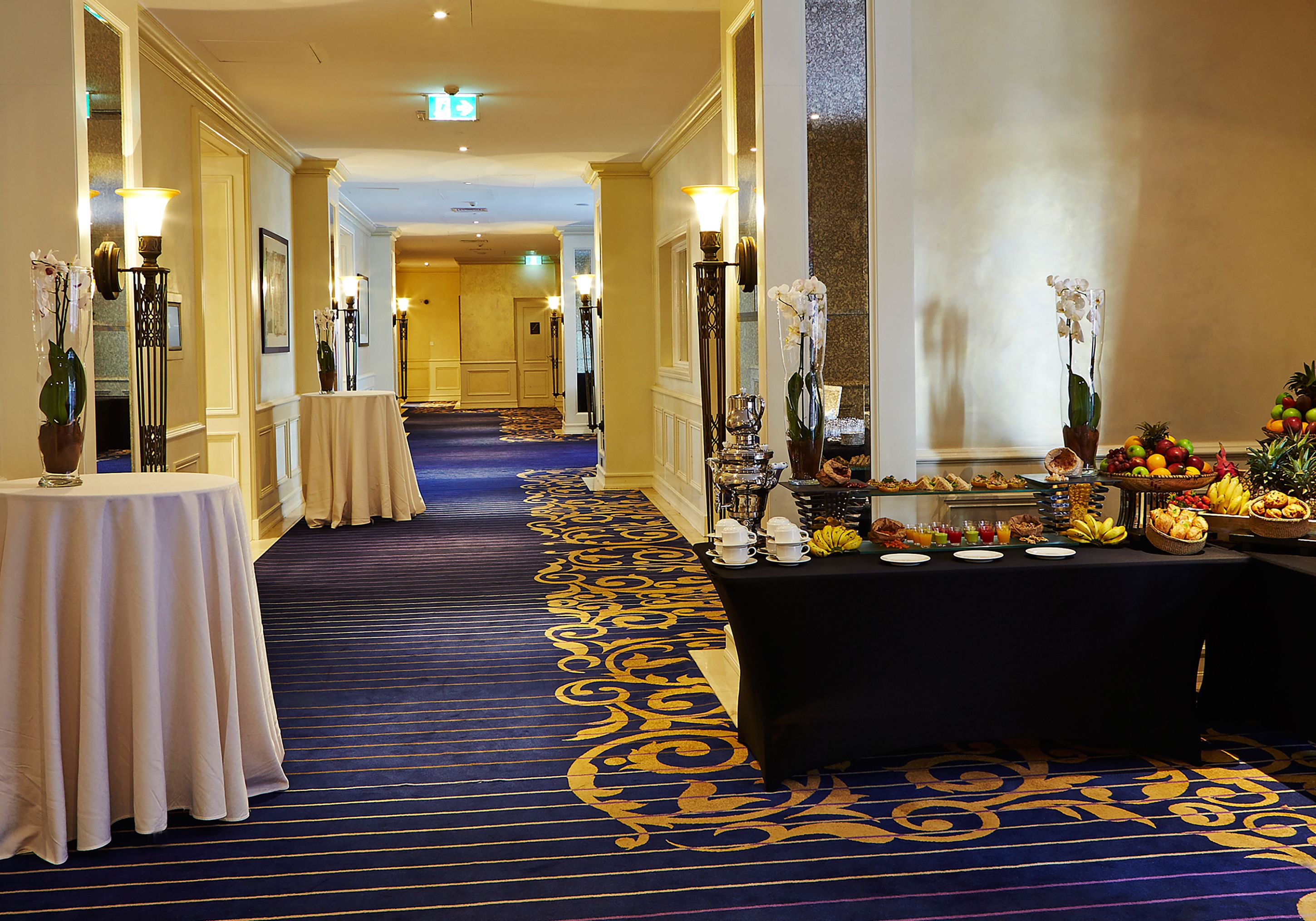A hotel corridor with a blue carpet, tables with white tablecloths, and a buffet table with fruits and beverages.