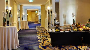 A hotel corridor with a blue carpet, tables with white tablecloths, and a buffet table with fruits and beverages.