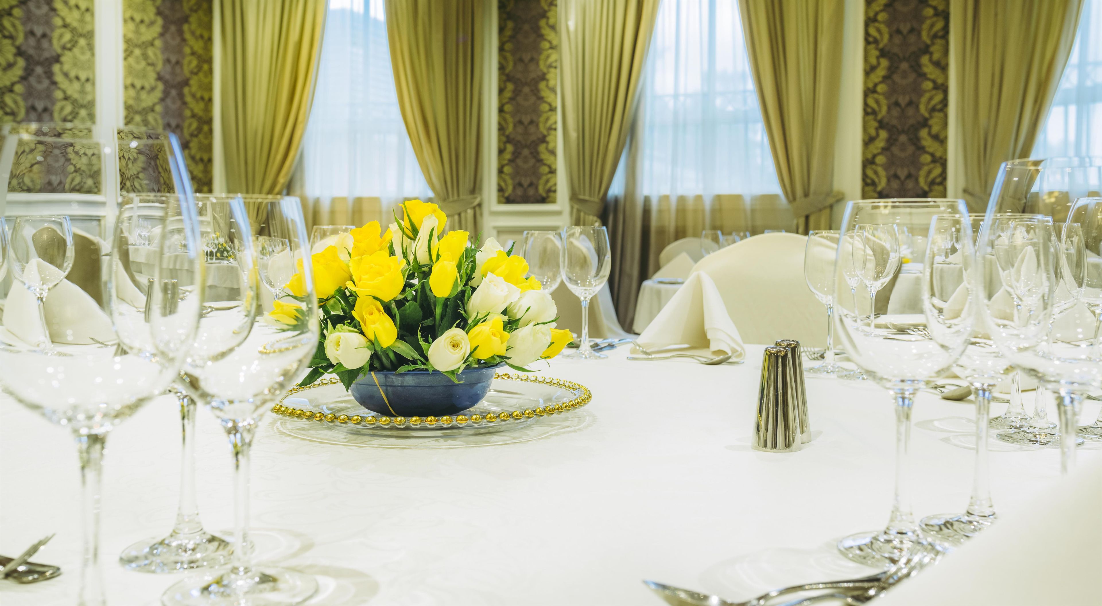 A formal dining table set with multiple wine glasses, a centerpiece of yellow and white flowers, and a gold-trimmed charger plate.