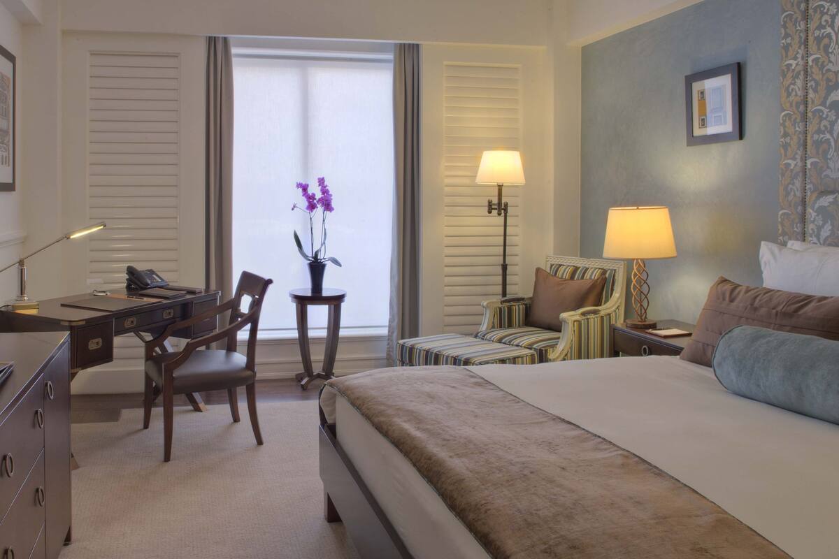 A hotel room with a bed, desk, chairs, lamps, and a window with curtains.