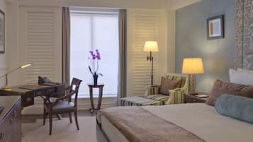 A hotel room with a bed, desk, chairs, lamps, and a window with curtains.