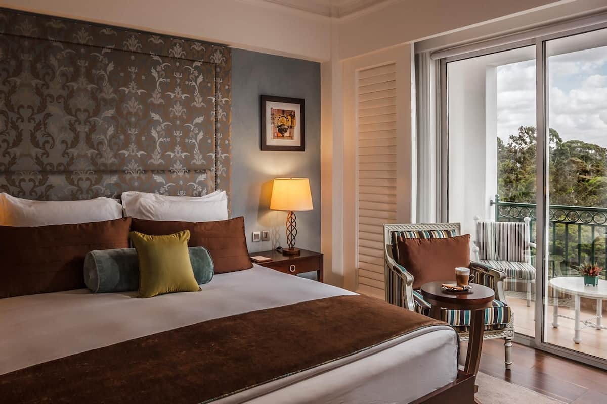 A hotel room with a large bed, a nightstand with a lamp, a chair, and a balcony with a table and chairs.