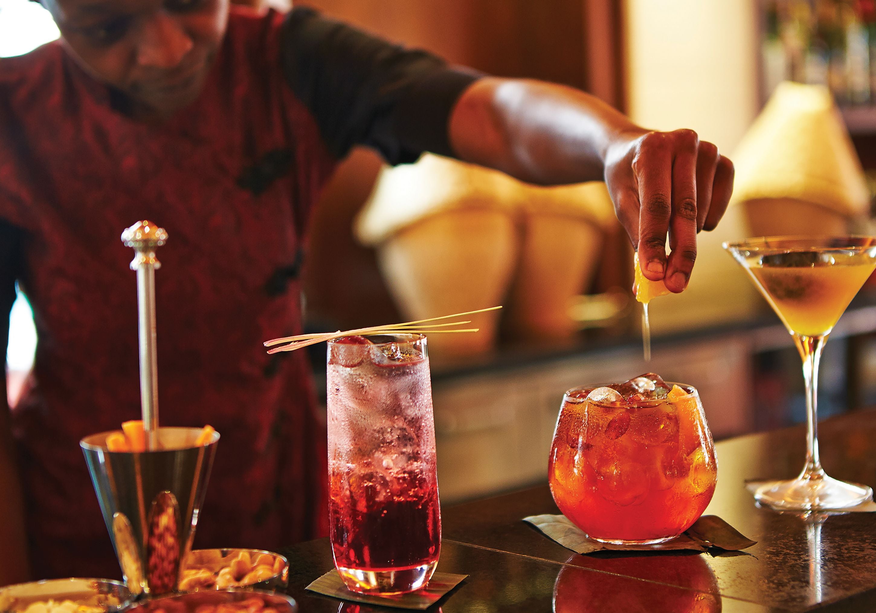 Un bar con una persona che versa un drink in un bicchiere, una coppa Martini e un bicchiere alto con una cannuccia.