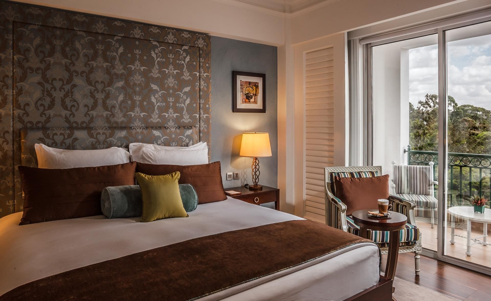 A hotel room with a large bed, a nightstand with a lamp, a chair, and a balcony with a table and chairs.
