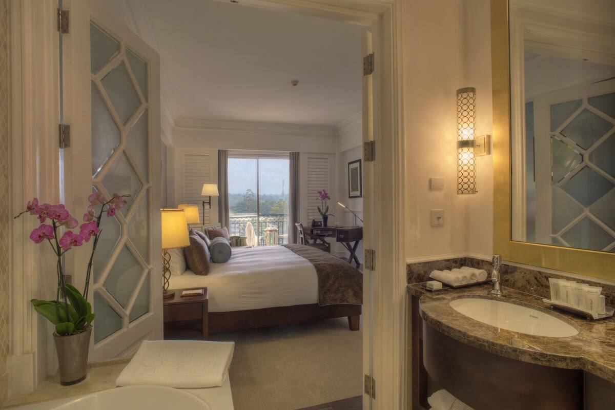 A hotel room with a large bed, a sitting area with a view, a bathroom with a sink and mirror, and a potted orchid.