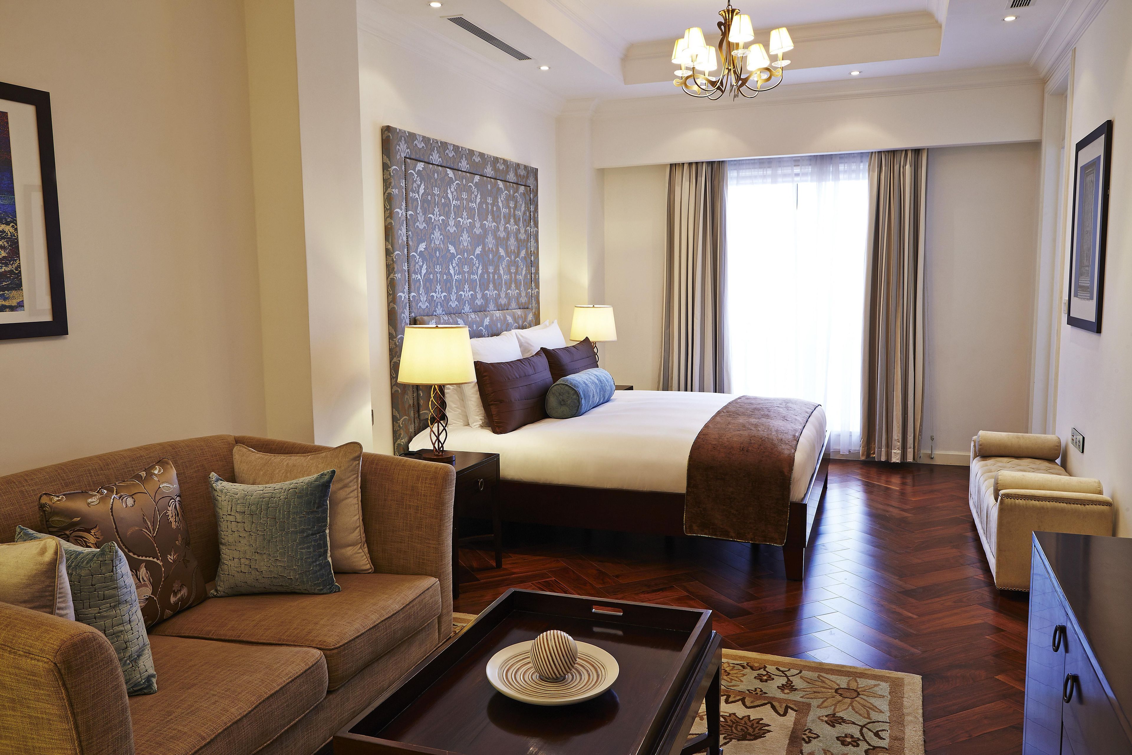 A hotel room with a large bed, a sofa, a coffee table with a hat, and a chandelier.