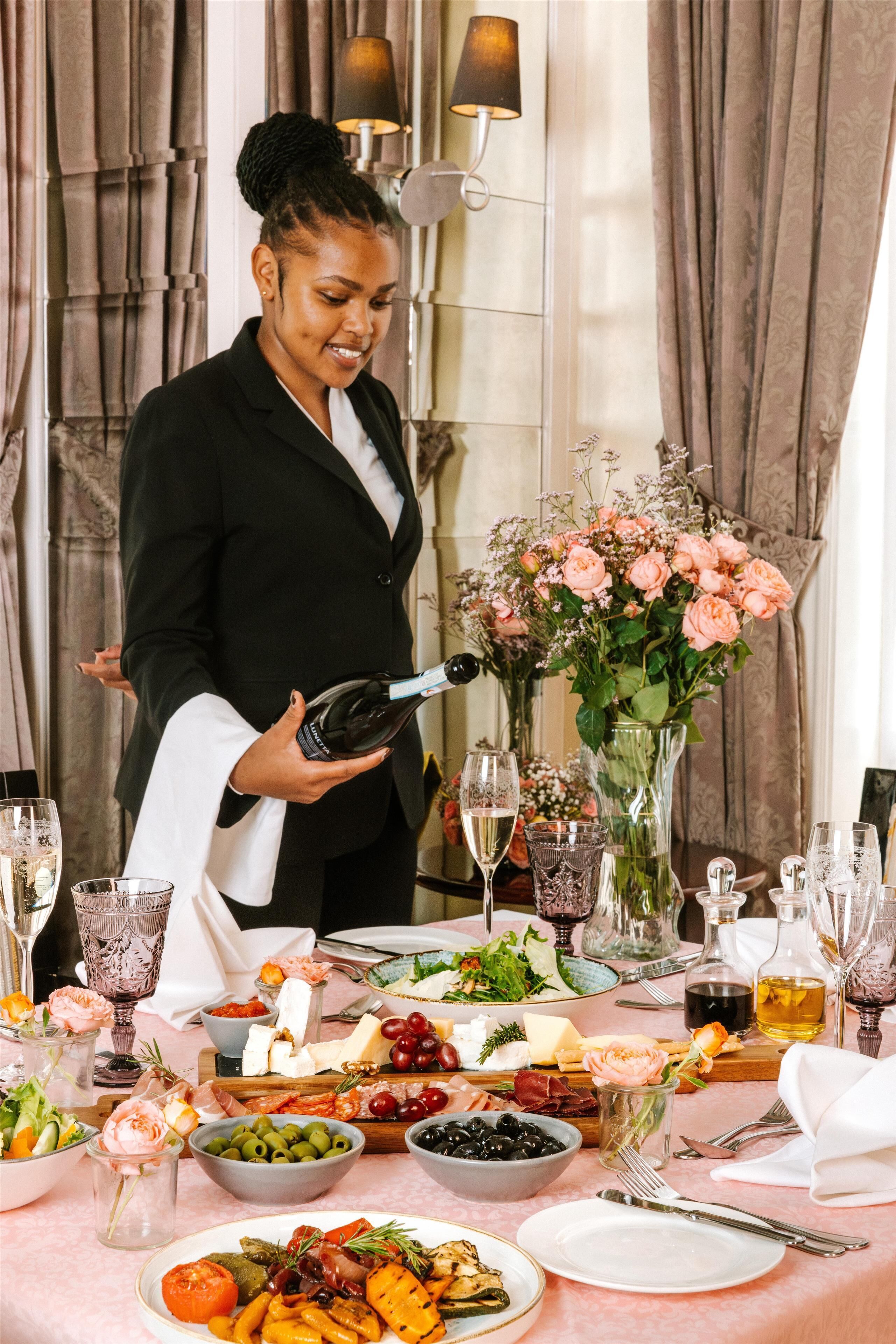 Disposição formal para refeição com uma mesa posta, uma pessoa segurando uma garrafa e um vaso com flores.