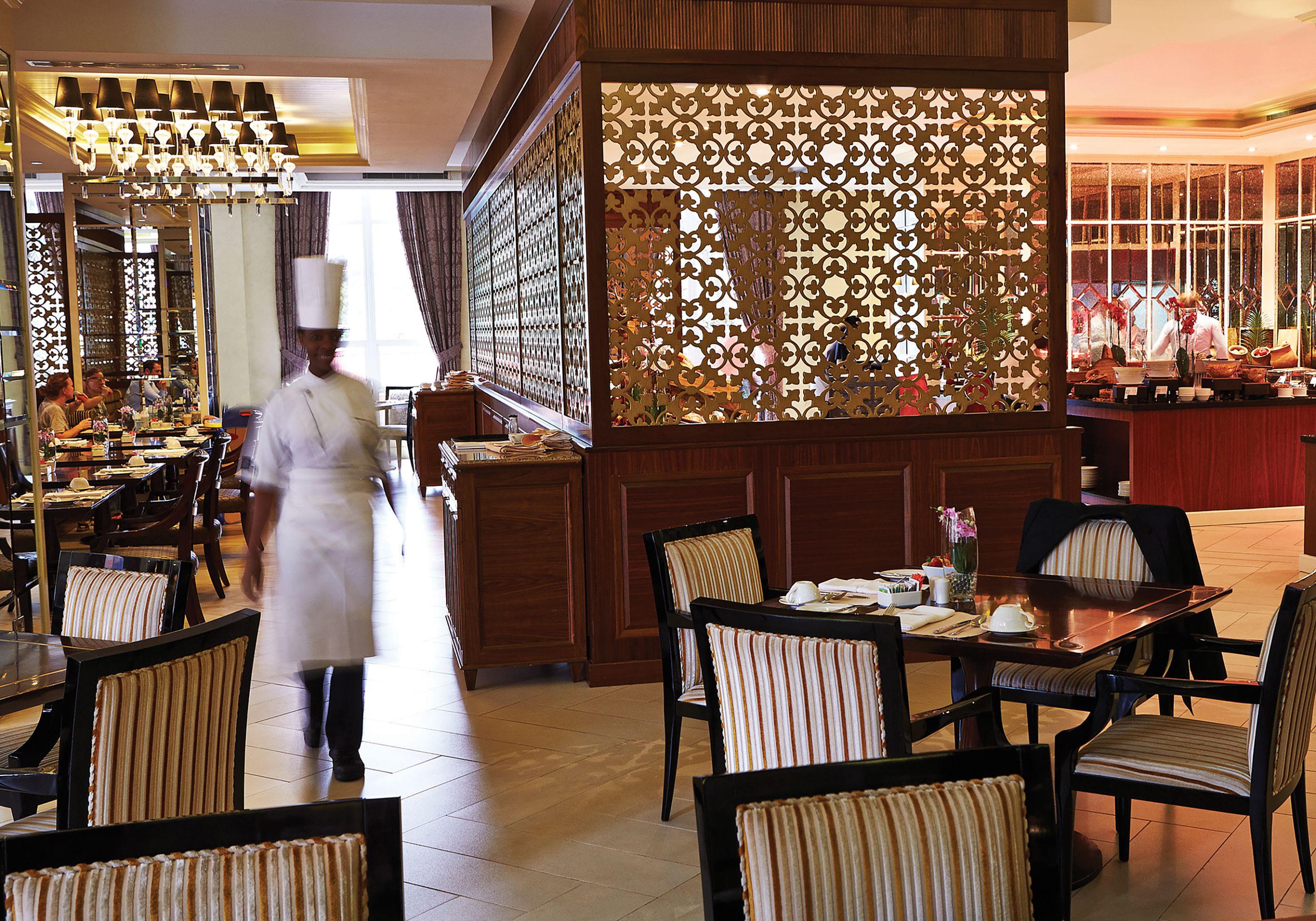 Un restaurante con un chef caminando, muebles de madera y un separador decorativo.