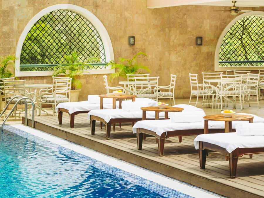 A poolside area with lounge chairs and tables, a pool, and arched windows with greenery visible through the mesh.