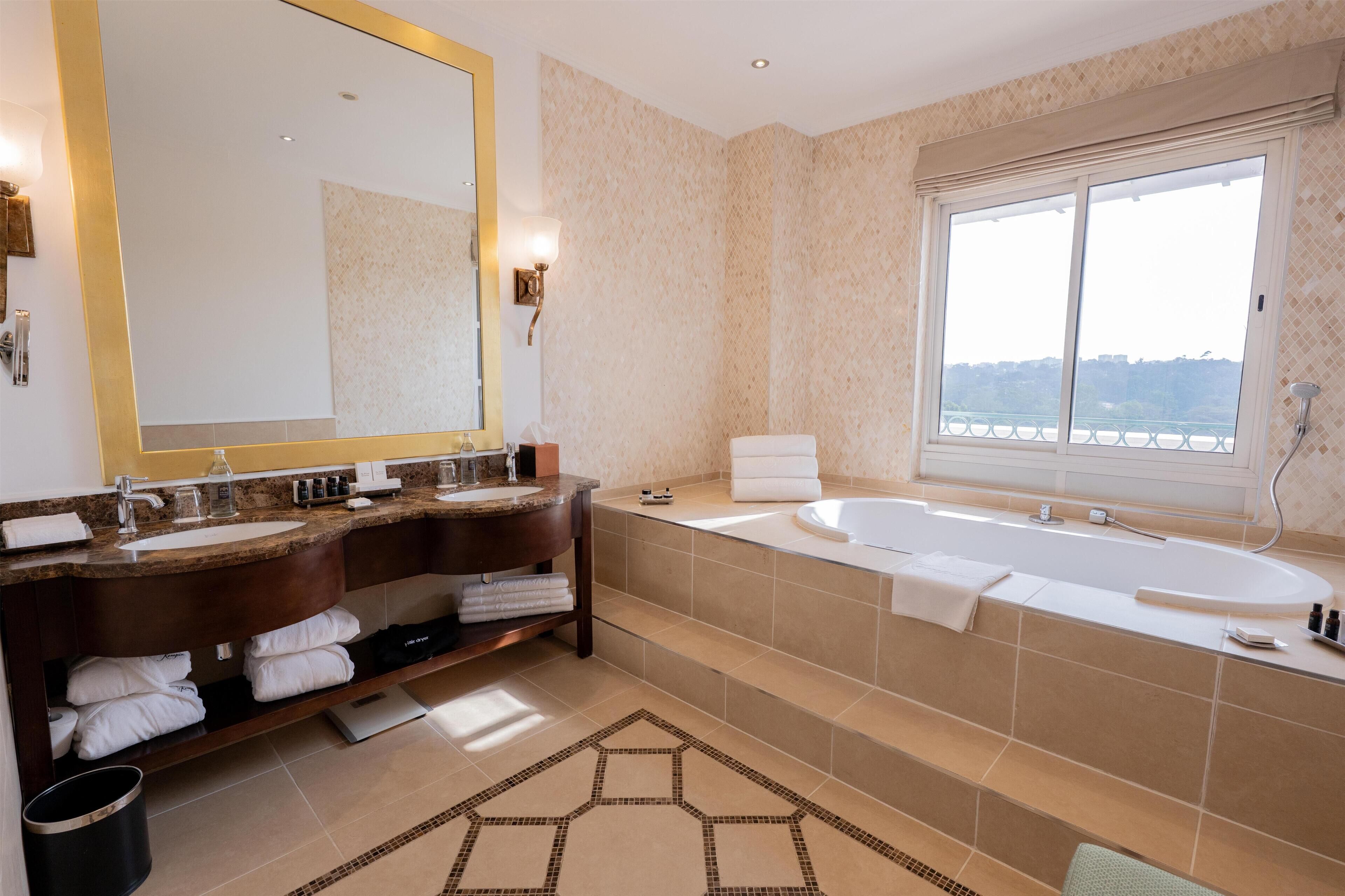 A modern bathroom with a large mirror, dual sinks, a bathtub, and a view through a large window.