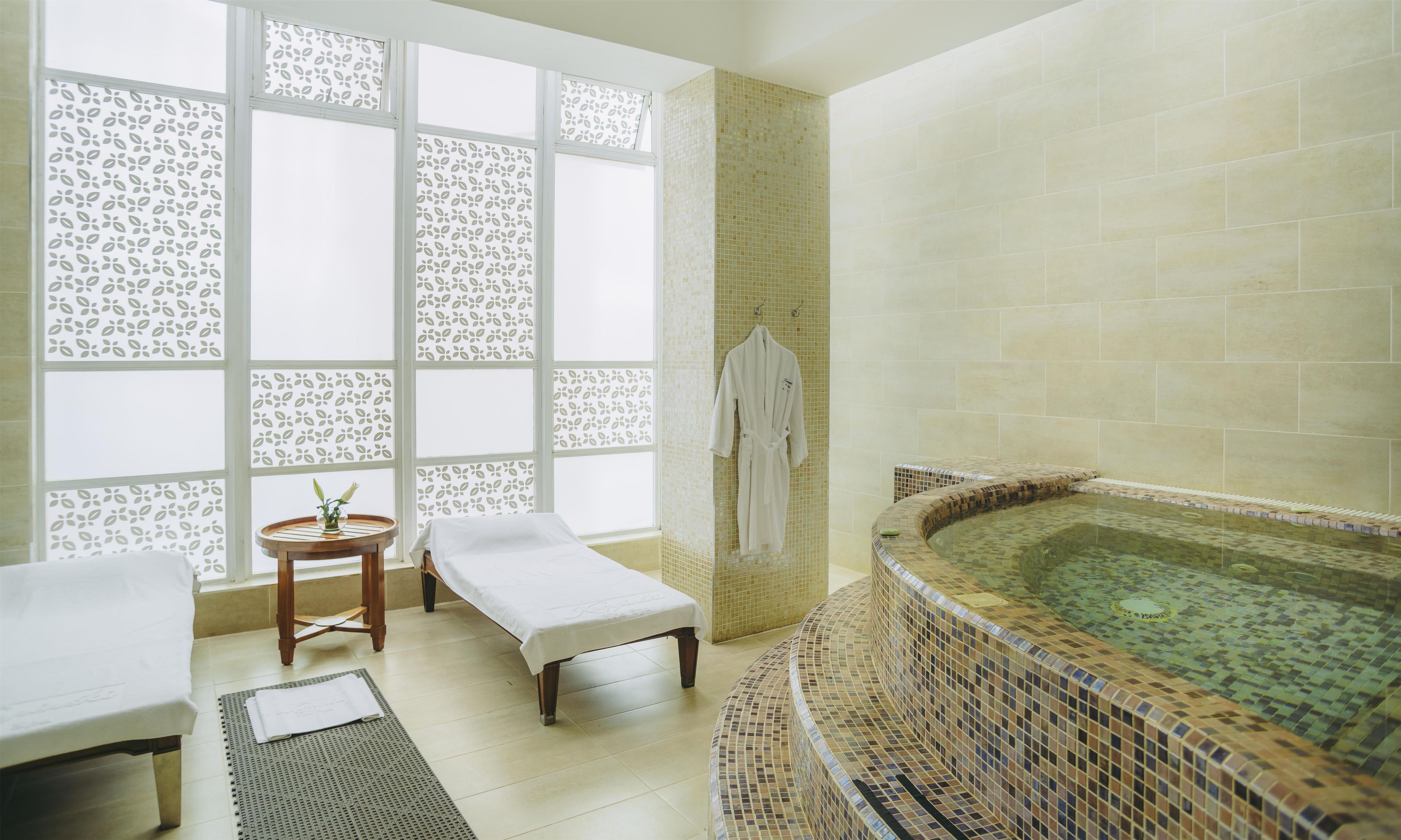 Una sala de spa con un ventanal grande, una camilla de masaje, una mesita con un florero y una bañera de hidromasaje.