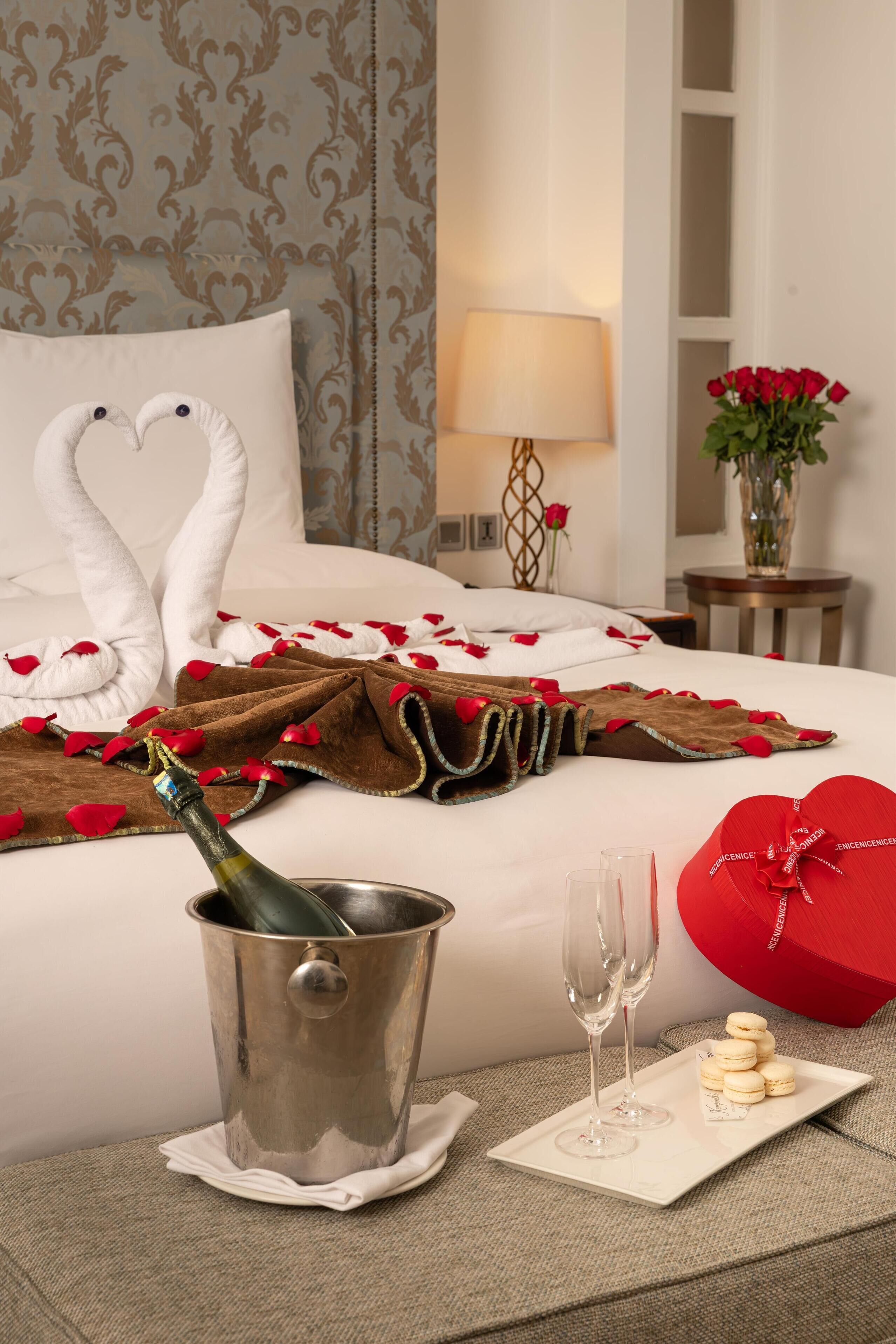 A neatly made bed with heart-shaped towels, a bottle of champagne, two glasses, and a tray with macarons.