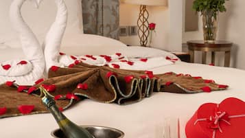 A neatly made bed with heart-shaped towels, a bottle of champagne, two glasses, and a tray with macarons.