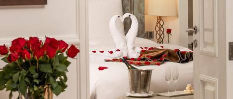 Una habitación de hotel con cama, una mesita de noche con un jarrón de rosas rojas, una lámpara de noche y un panel decorativo en la pared.