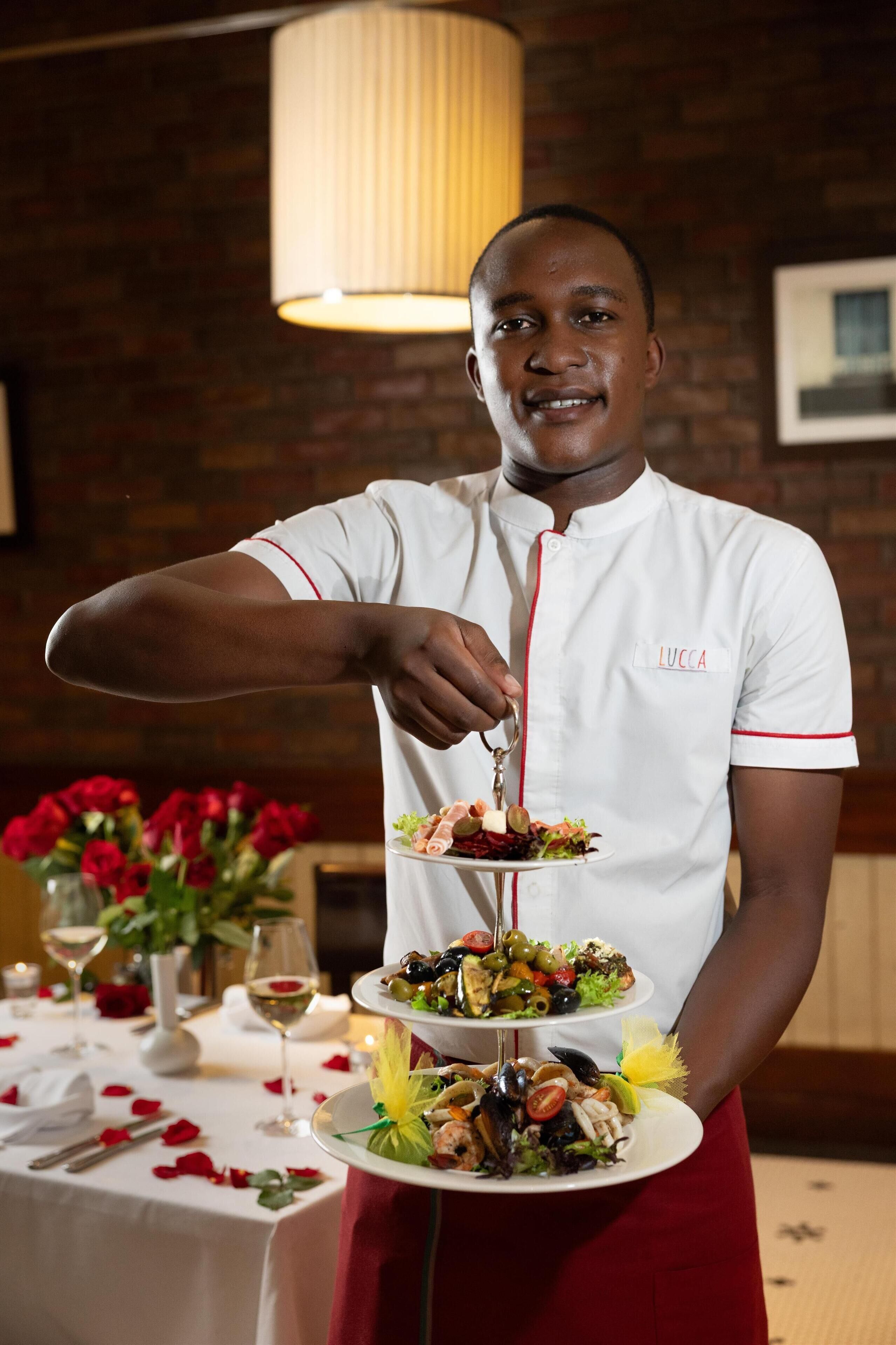 A person in a white chef's uniform with red trim, holding a three-tiered plate of food.