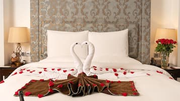 A neatly made bed with a heart-shaped pillow and two swan-shaped pillows, a champagne bucket, two glasses, a heart-shaped box, and a bouquet of roses on a table.