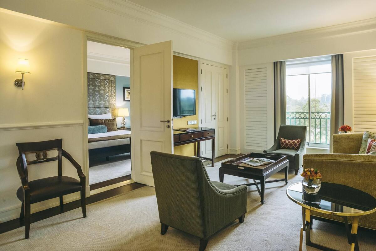 A hotel room with a bed, a desk with a TV, a sofa, a chair, and a balcony with a view.