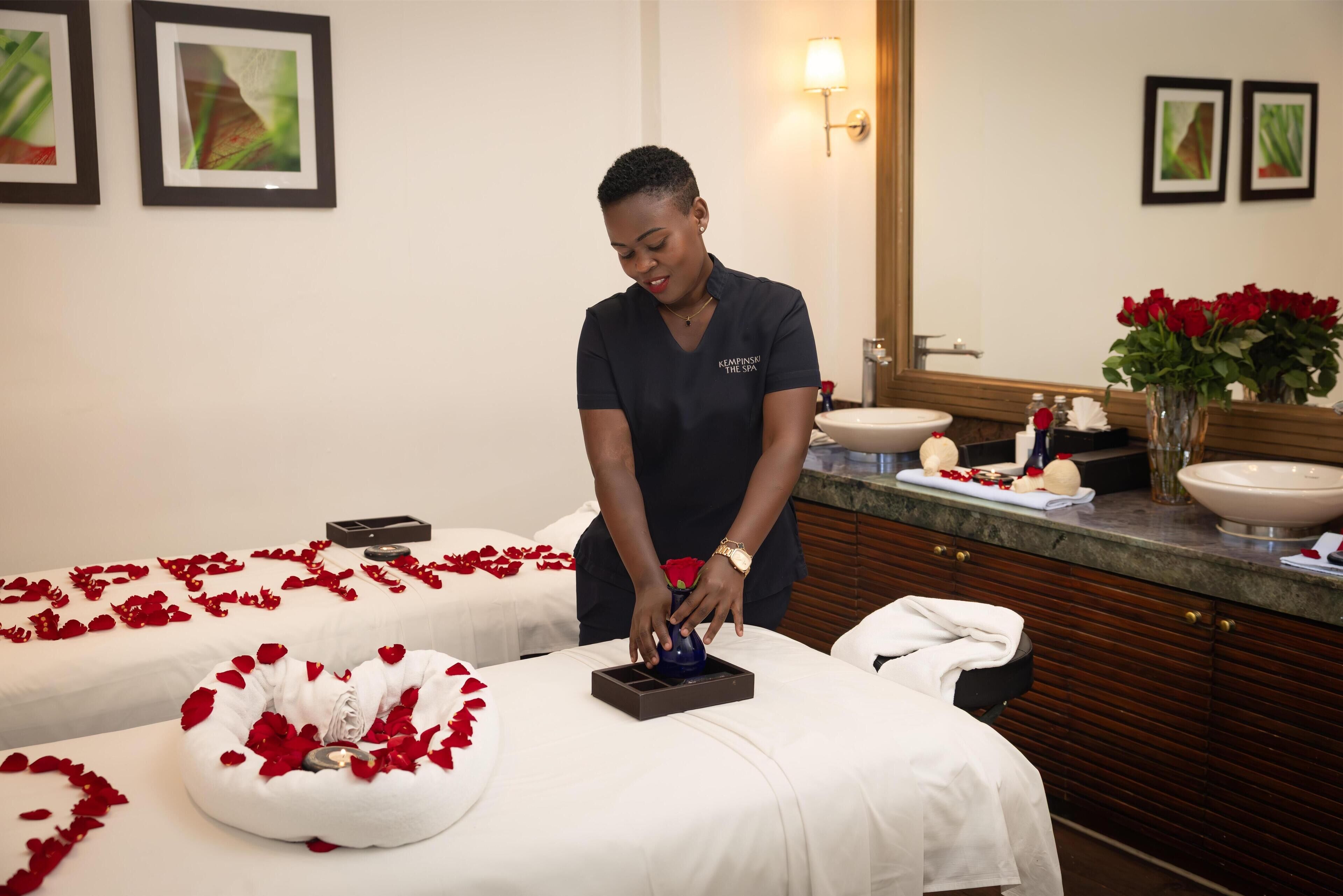 Une salle de spa équipée de deux tables de massage recouvertes de draps blancs et de pétales de roses rouges, d’un lavabo et d’un miroir.