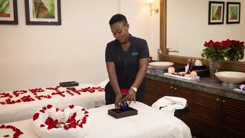 Une salle de spa équipée de deux tables de massage recouvertes de draps blancs et de pétales de roses rouges, d’un lavabo et d’un miroir.