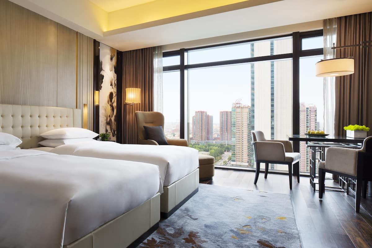 A hotel room with two beds, a sitting area with a table and chairs, a large window with a city view, and a modern design.