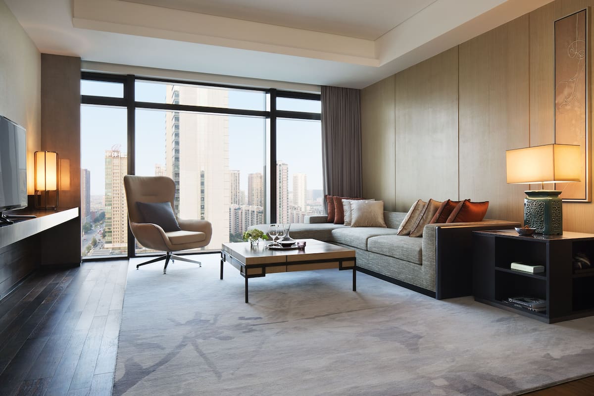 A modern living room with a sofa, armchair, and coffee table, large windows with a city view, and a TV on a stand.