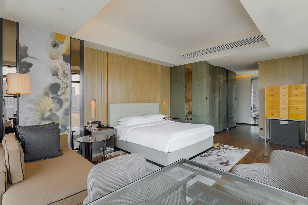 A modern hotel room with a large bed, a sofa, a glass table, and a view of the city through large windows.