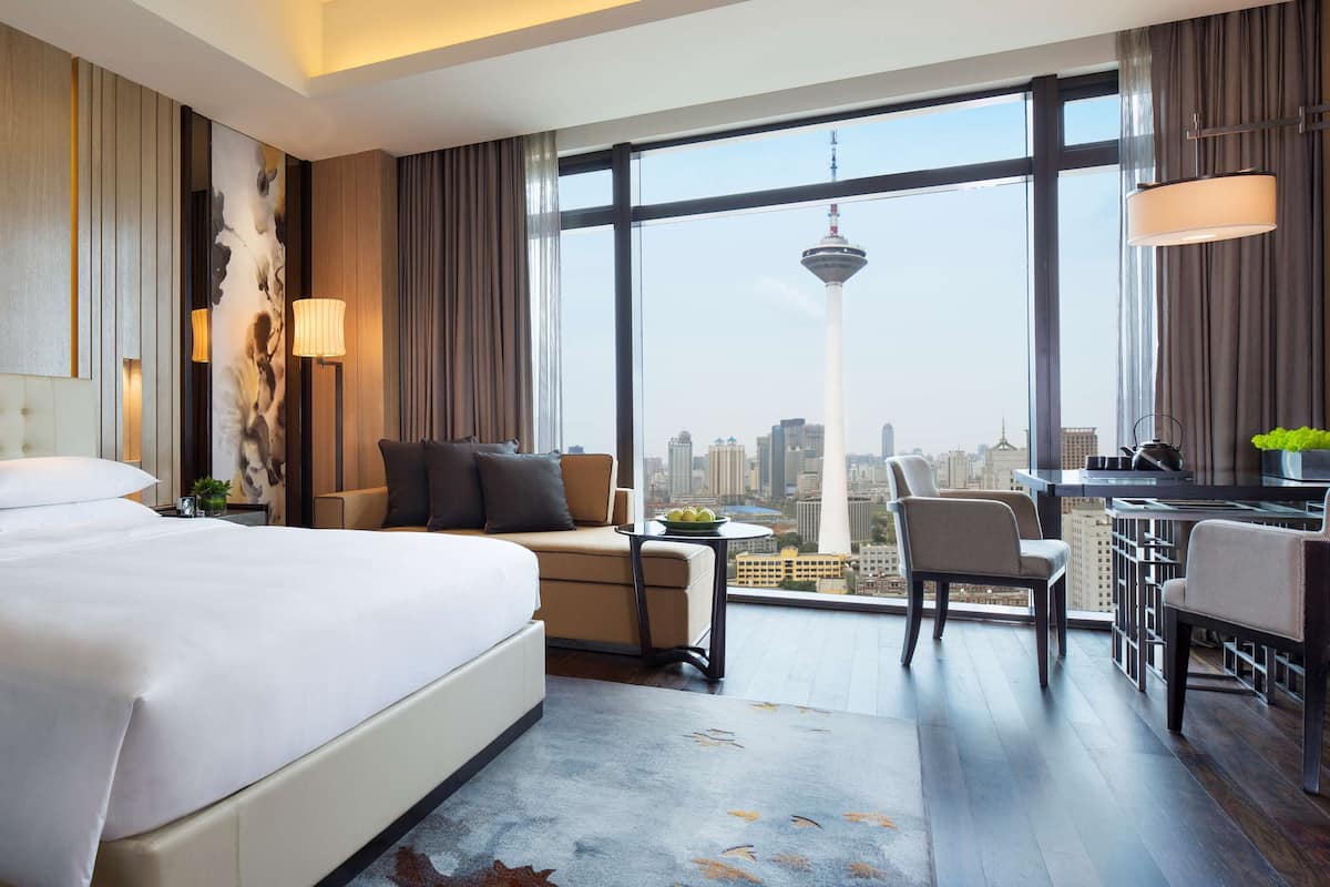 A modern hotel room with a large bed, a seating area with a view of the city, and a dining area.