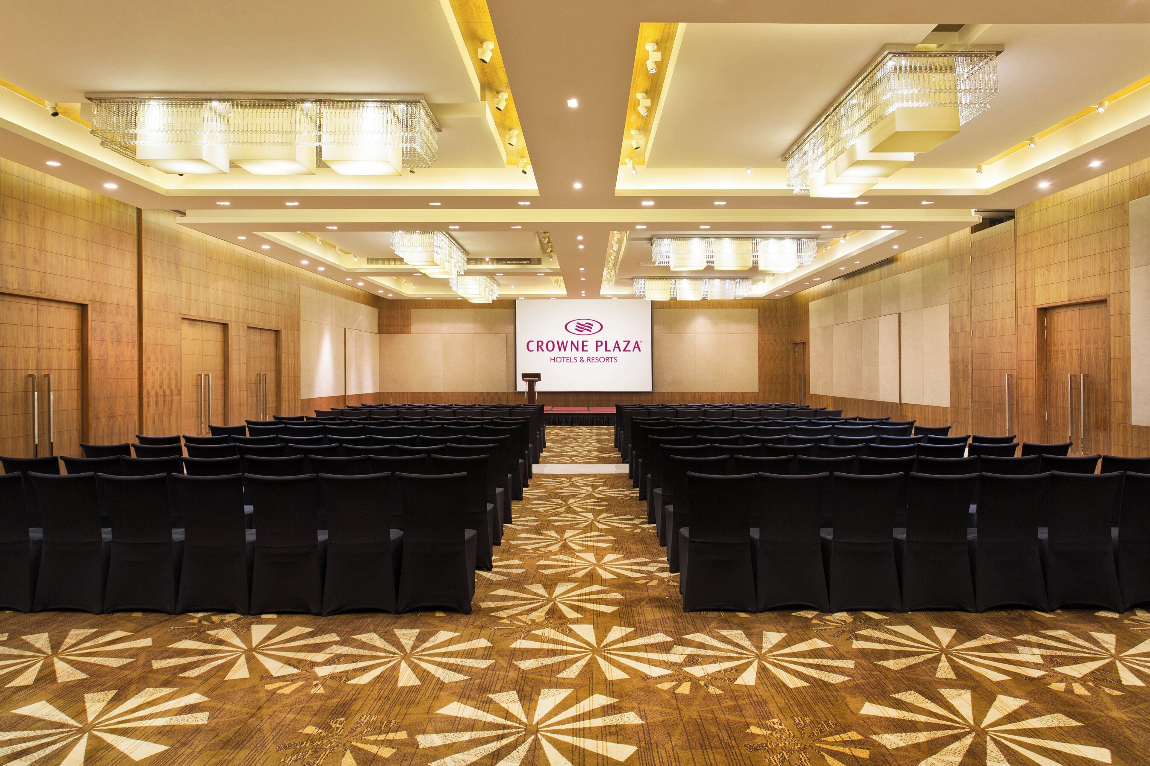 A conference room with rows of chairs, a podium, and a banner reading 'CROWN PLAZA HOTELS & RESORTS'.