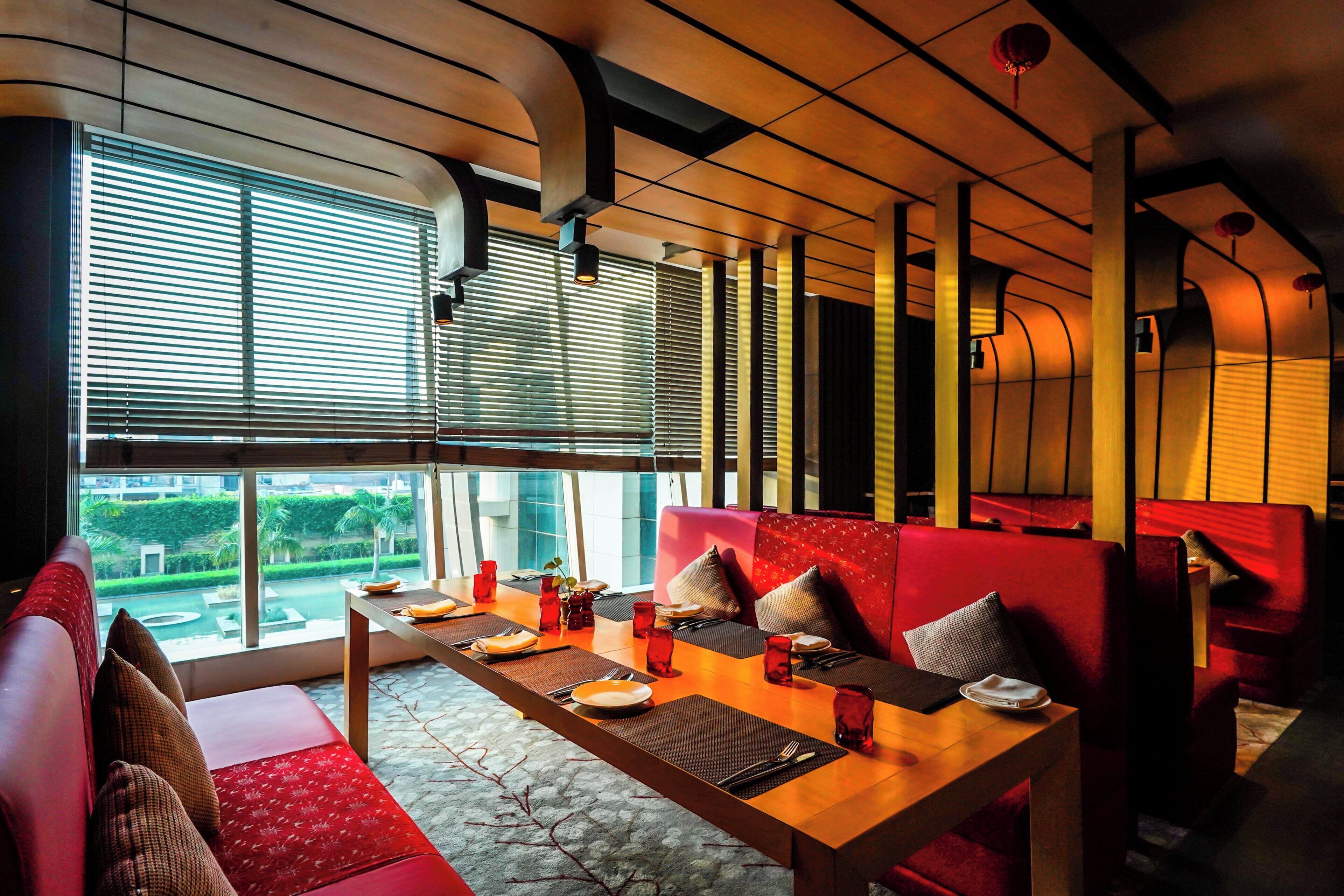 A dining area with a table set for two, red upholstered seating, and large windows with blinds.