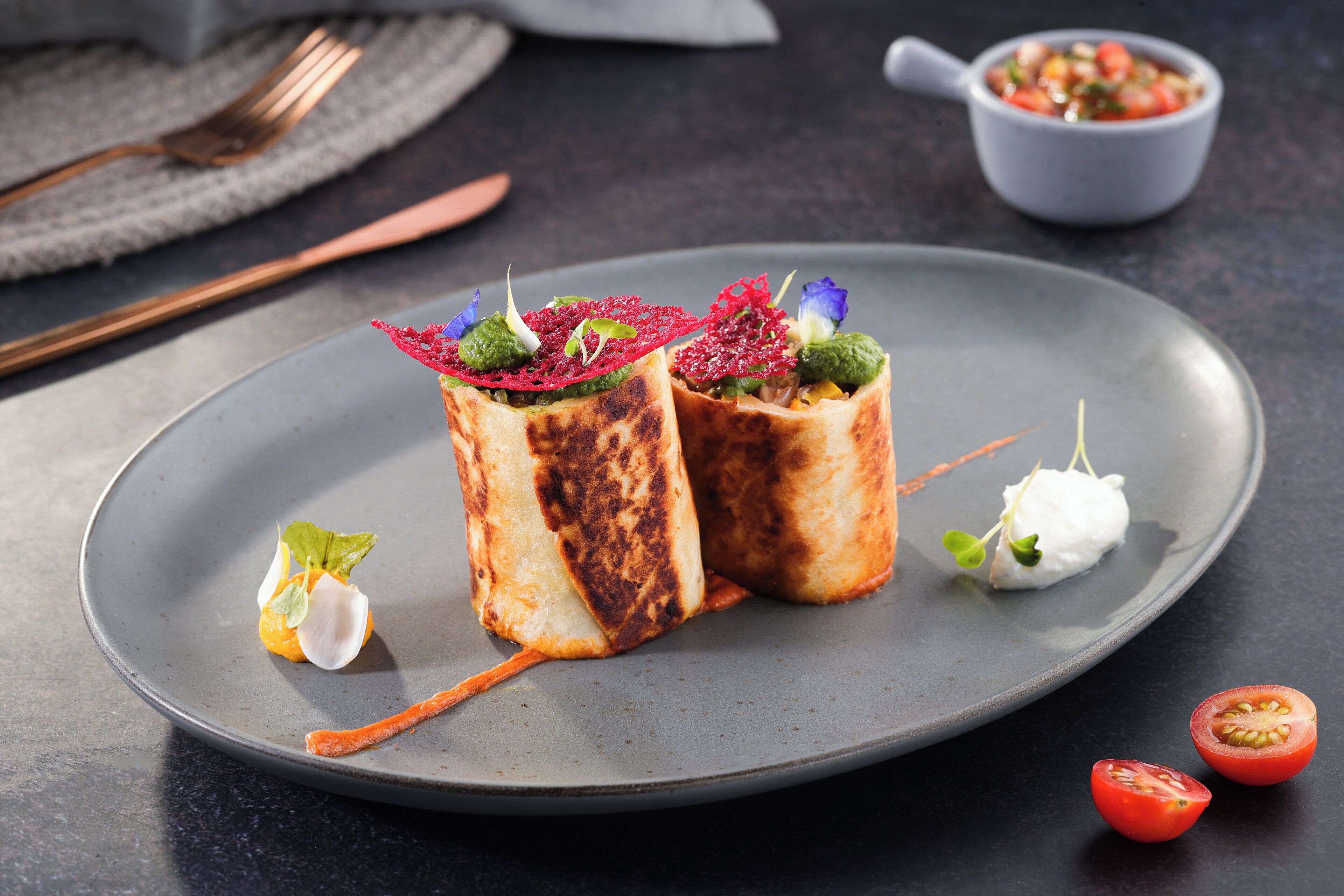 Un plato con dos porciones envueltas, decorado con vegetales y una pequeña cucharada blanca, y un recipiente con salsa al fondo.