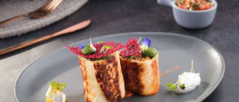 Un plato con dos porciones envueltas, decorado con vegetales y una pequeña cucharada blanca, y un recipiente con salsa al fondo.