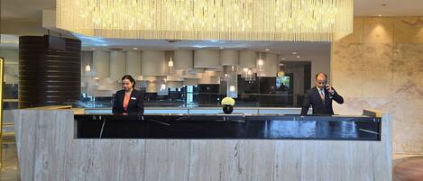 A reception desk with two staff members, a modern chandelier, and marble walls.