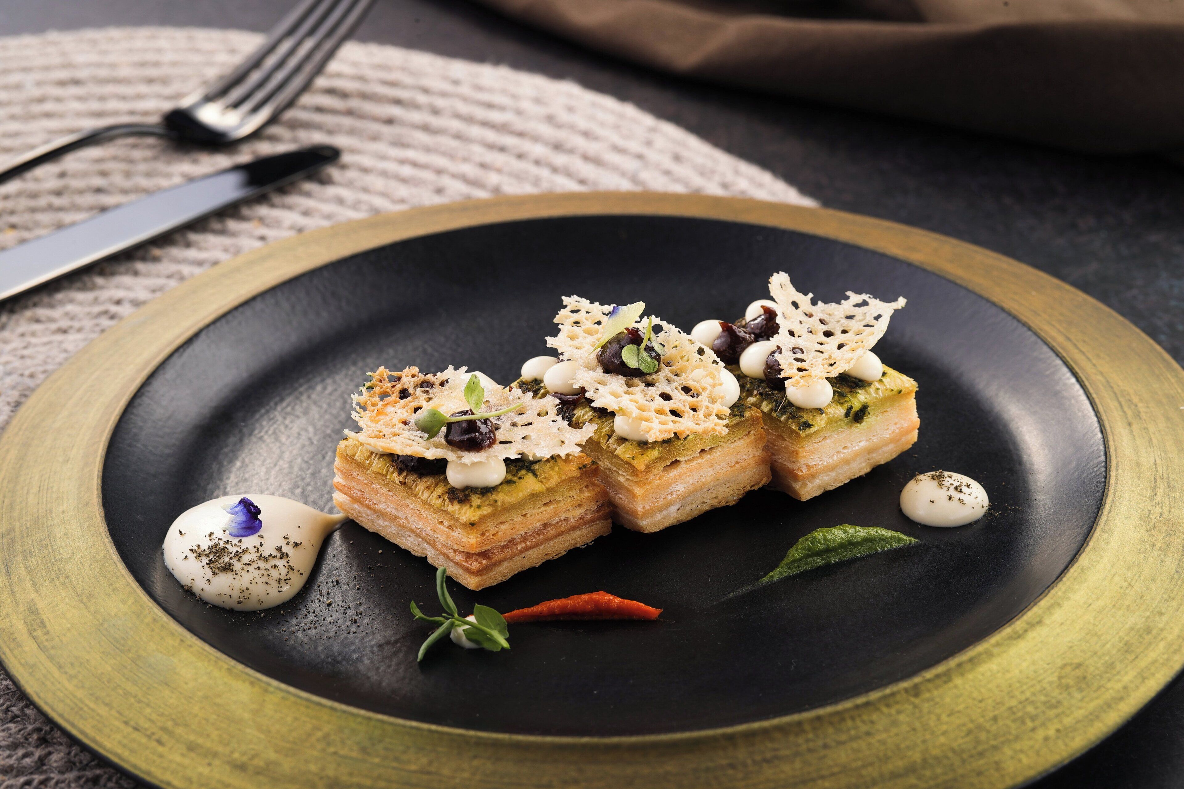Une assiette noire sur laquelle est présentée une pâtisserie feuilletée à plusieurs couches, décorée d’éléments blancs et verts, posée sur une nappe à motifs.
