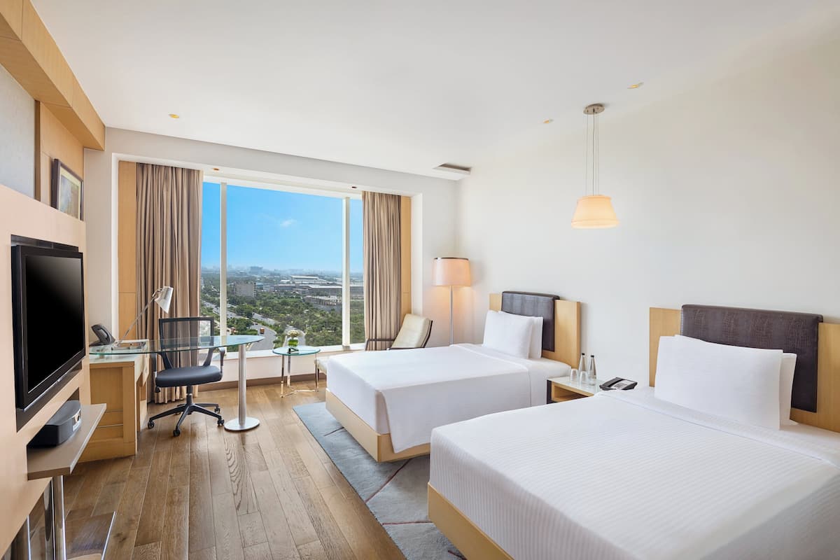 Habitación de hotel con dos camas, un escritorio con computadora, televisión y un ventanal con vista a la ciudad.